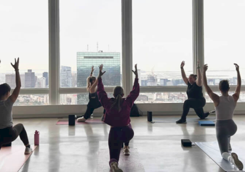 People doing yoga in a room with large windows, overlooking a cityscape. They are in a lunge pose with arms raised.