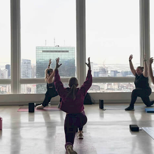 People doing yoga in a room with large windows, overlooking a cityscape. They are in a lunge pose with arms raised.