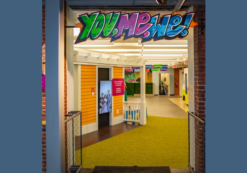 A brightly lit, colorful hallway with a sign that reads "YOU, ME, WE!" in graffiti-style lettering. There are yellow walls and green flooring.