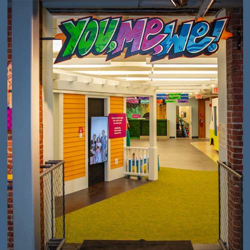 A brightly lit, colorful hallway with a sign that reads "YOU, ME, WE!" in graffiti-style lettering. There are yellow walls and green flooring.