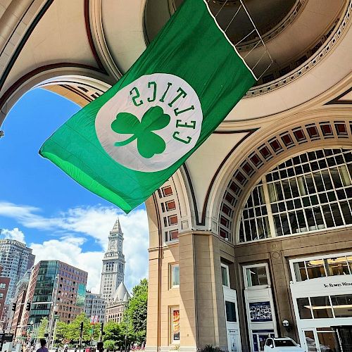 The image shows a green Celtics flag with a shamrock hanging under an arched structure, with a cityscape and clock tower in the background.