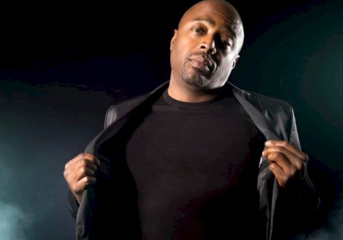 A man in a black outfit is posing confidently while holding his jacket open against a dark background.