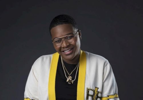 The image shows a person wearing glasses, a black shirt, and a white jacket with yellow trim, smiling at the camera against a dark background.