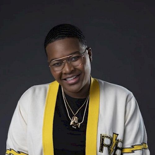 The image shows a person wearing glasses, a black shirt, and a white jacket with yellow trim, smiling at the camera against a dark background.