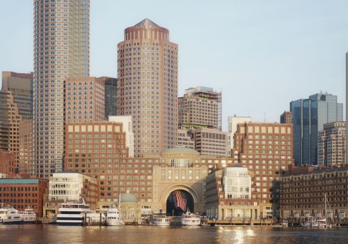 The image shows a waterfront cityscape with a mix of modern and classic buildings, several yachts docked at the harbor, and sunny weather.