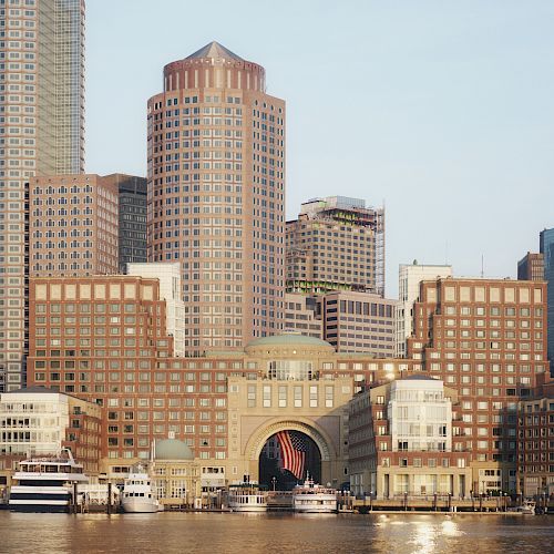 The image shows a waterfront cityscape with a mix of modern and classic buildings, several yachts docked at the harbor, and sunny weather.