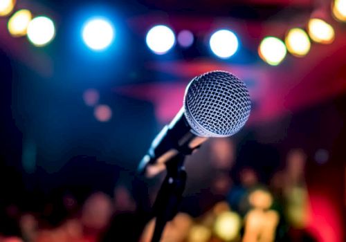 A microphone in the foreground is set up on a stage with colorful, blurred lights in the background, suggesting a performance setting.