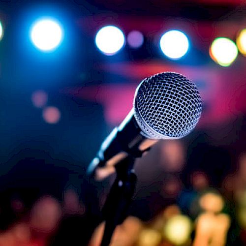 A close-up of a microphone on a stand, with colorful stage lights in the background, creating an atmosphere of a live performance.