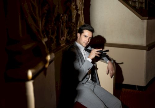 A man in a grey suit sits on a chair in a dimly lit room, holding a black object while looking at the camera.