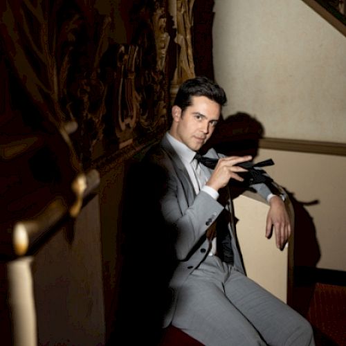 A man in a grey suit sits on a chair in a dimly lit room, holding a black object while looking at the camera.