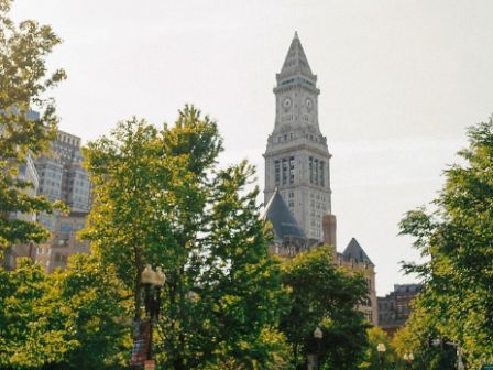 The image shows a tall clock tower building surrounded by trees and other buildings in an urban setting, likely a historical or notable landmark.