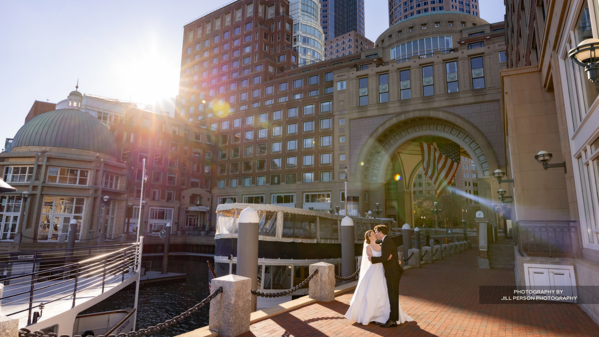 A newlywed couple poses for a wedding photo outdoors in a cityscape with sunlight streaming through buildings, an American flag, and waterfront.