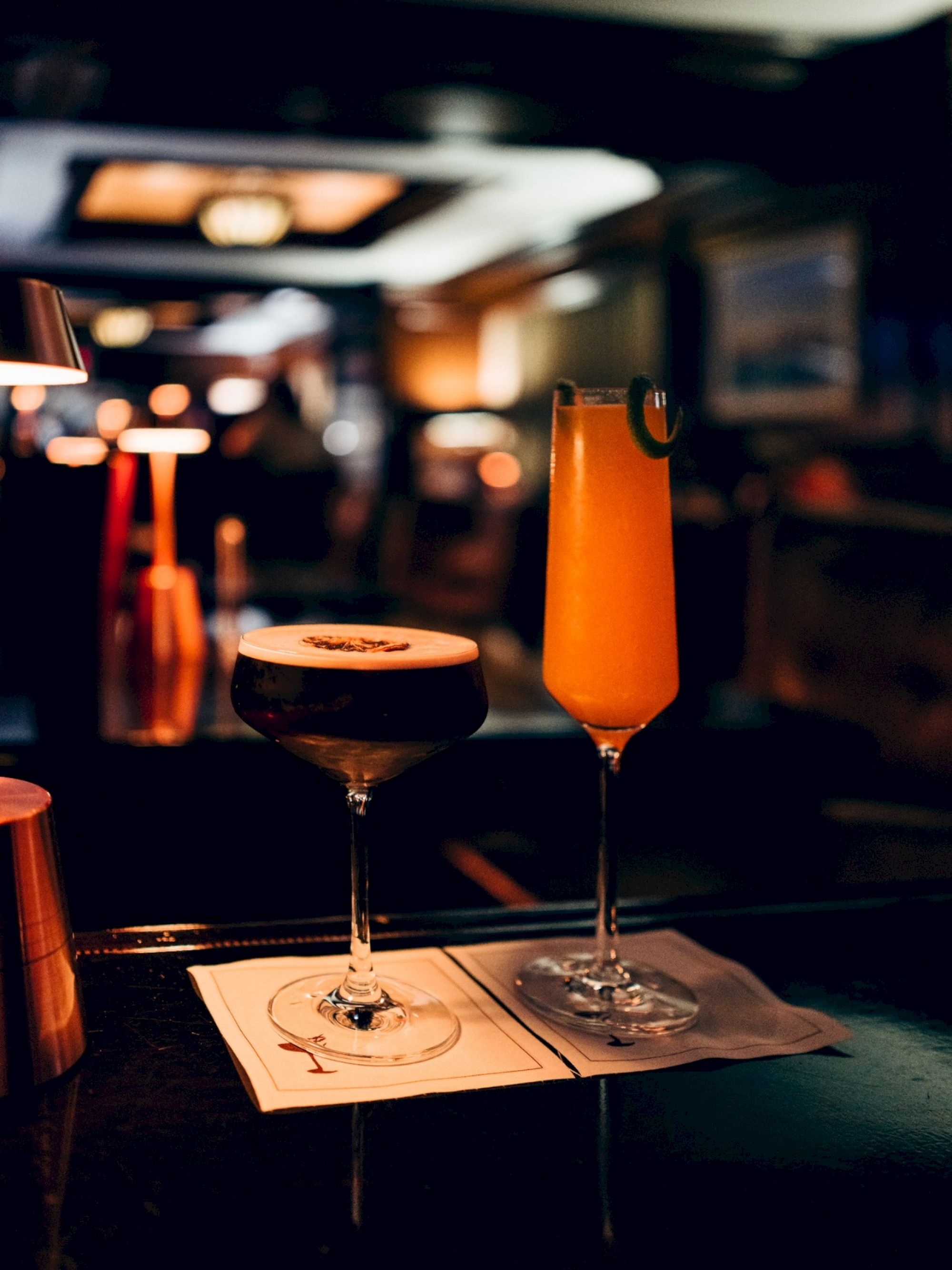 Two cocktails on a dimly lit bar counter, one dark with foam, the other orange with a garnish, on napkins.