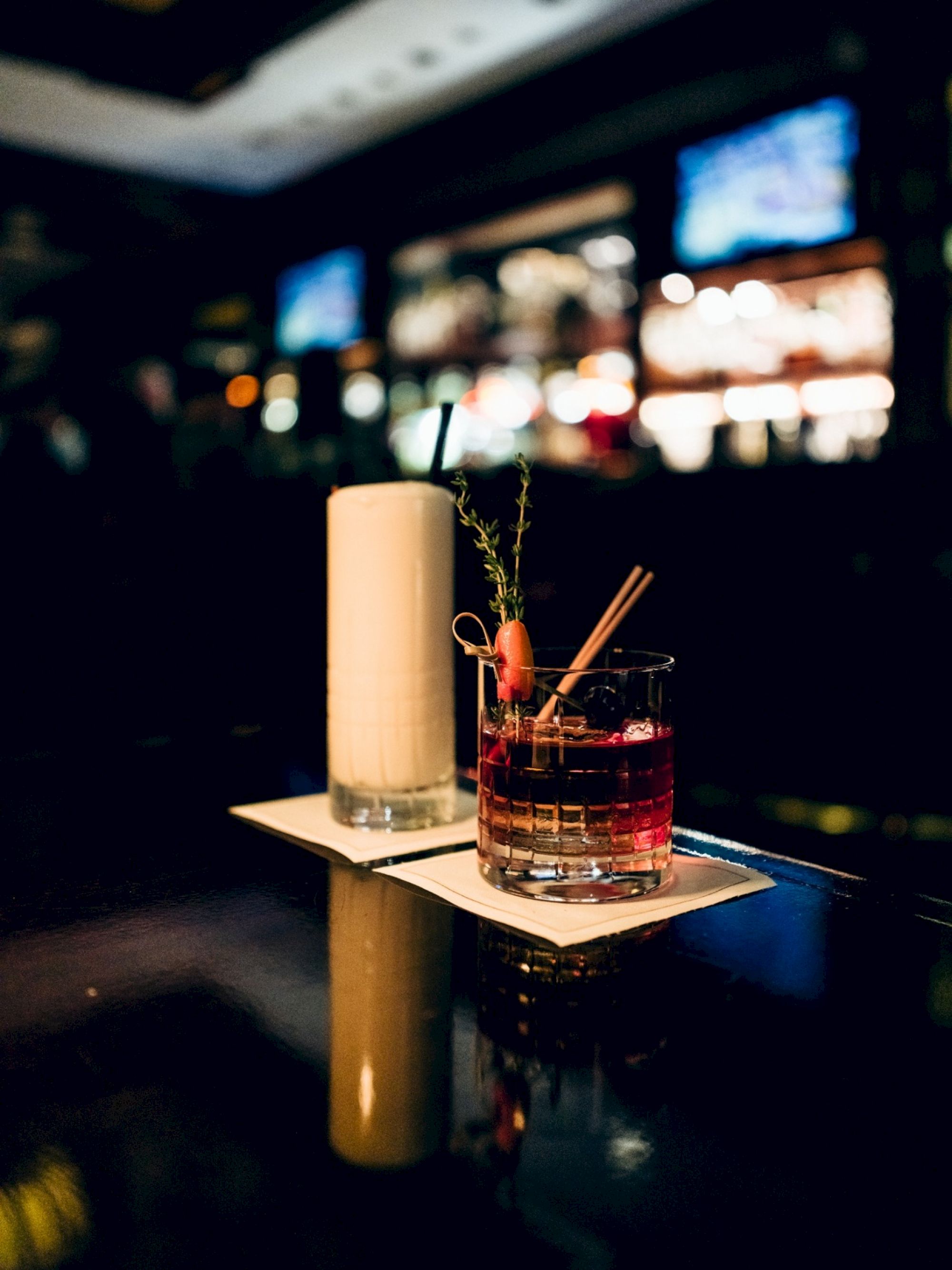 Two cocktails on a dark bar counter, one creamy and tall, the other reddish with garnish, in a dimly lit bar setting.