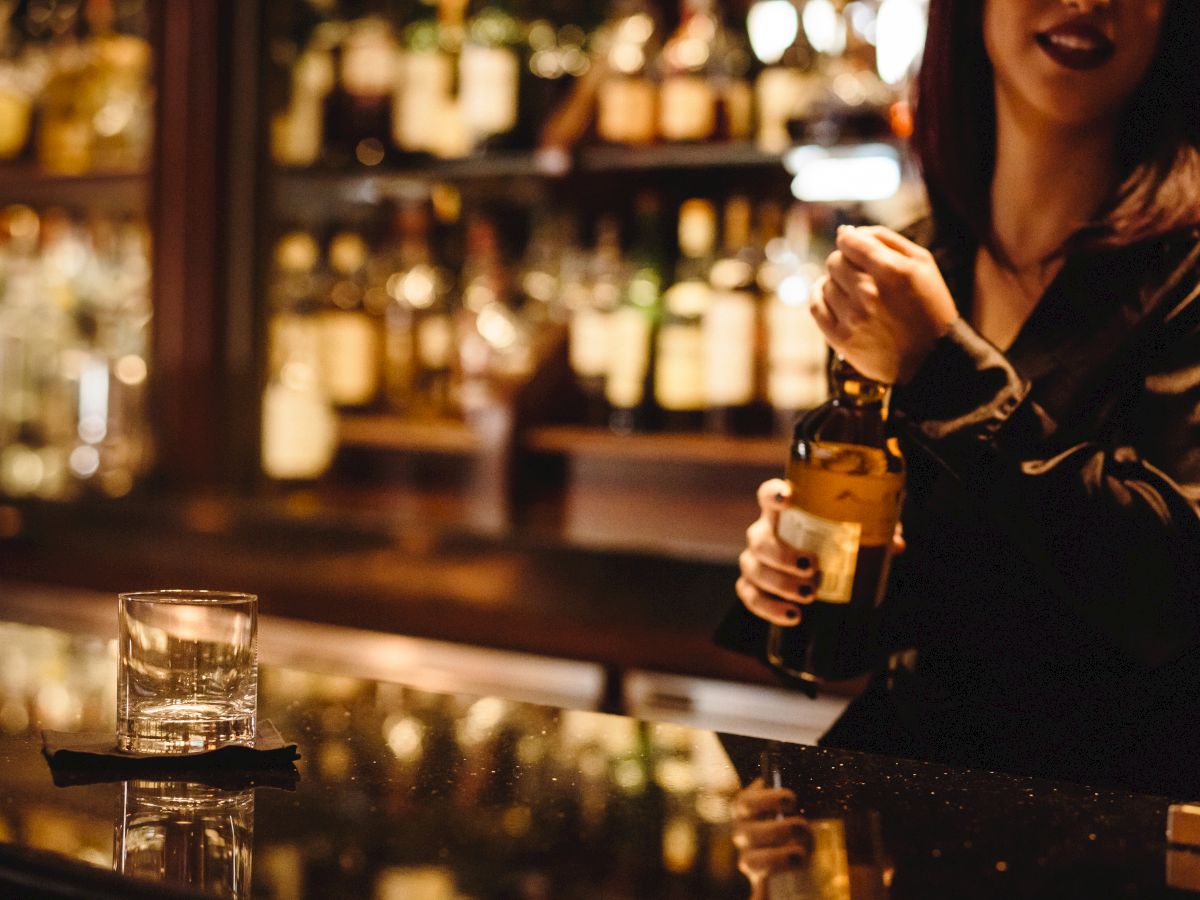 A bartender is holding a bottle of liquor behind a bar, with a glass on the countertop and various bottles in the background.