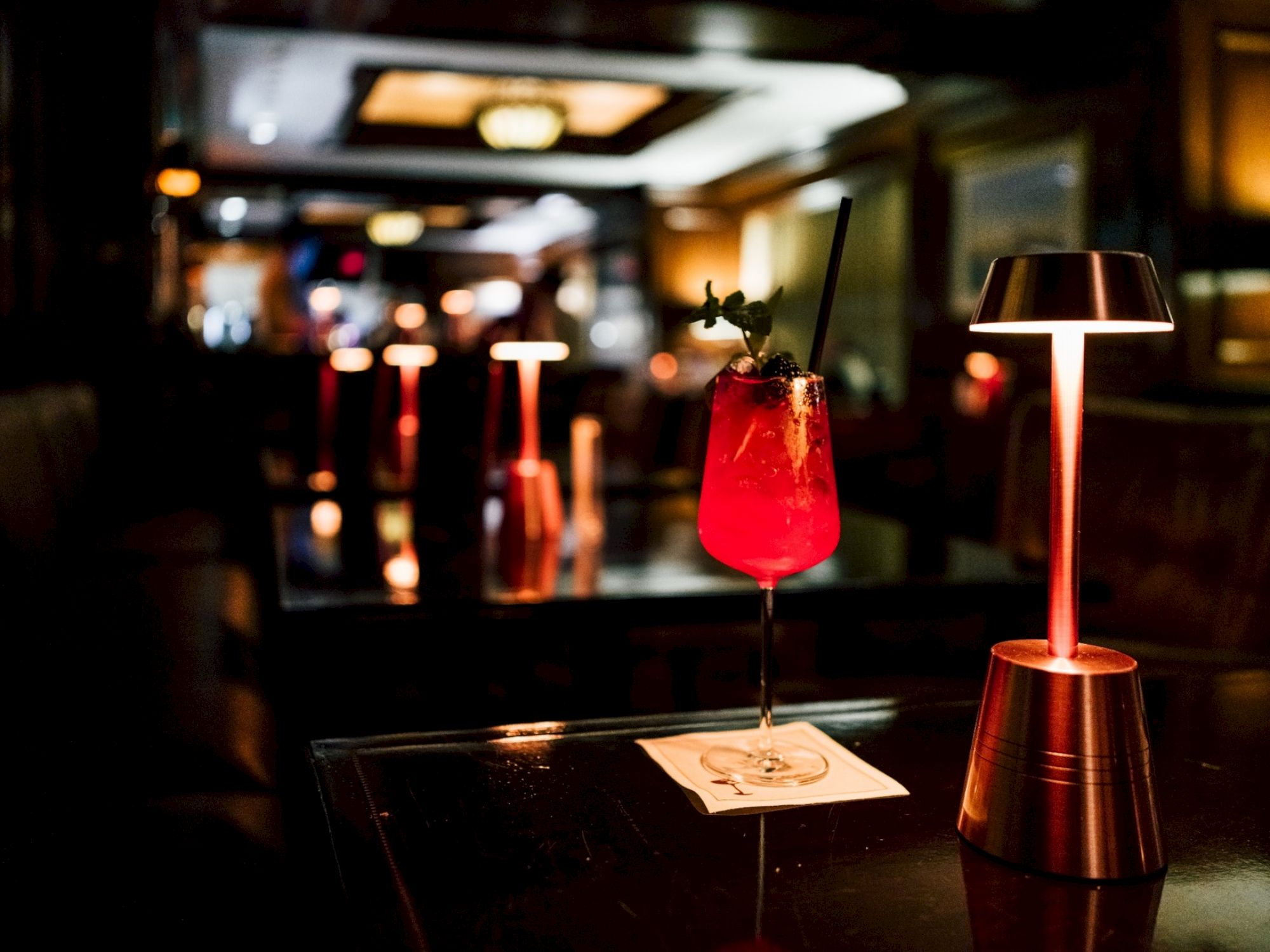 A dimly lit bar scene with a red cocktail on a coaster next to a stylish table lamp.