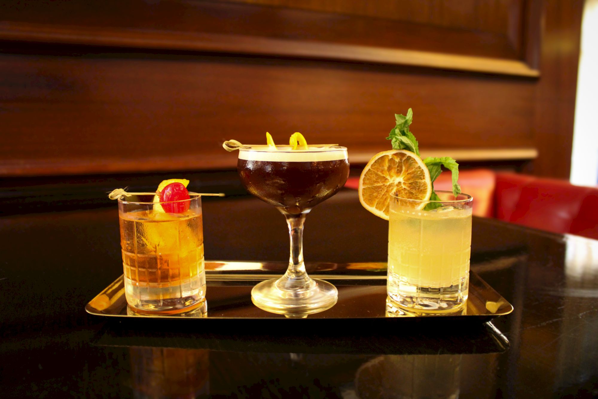 Three cocktails on a tray: one with an orange and cherry garnish, one in a coupe glass, and one with lemon and mint, all set against a wooden backdrop.