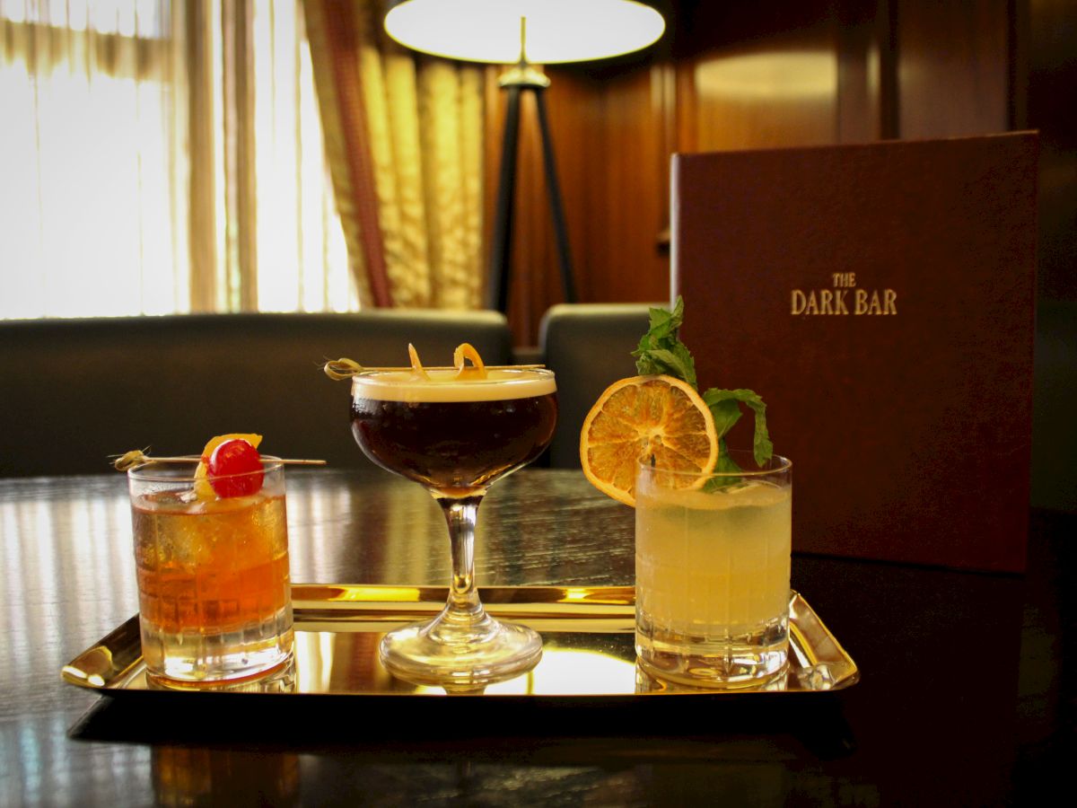 Three cocktails on a tray in a dimly lit bar, with a menu standing in the background and a lamp nearby, create an elegant setting.