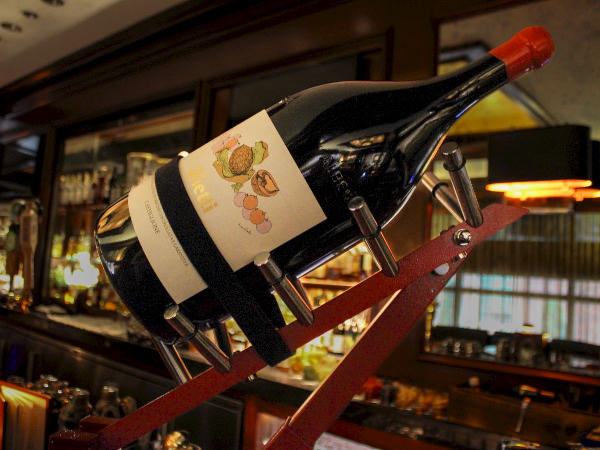 A large wine bottle displayed in a cradle at a bar, with a detailed label featuring grapes and other fruits, set in a dimly lit ambiance.