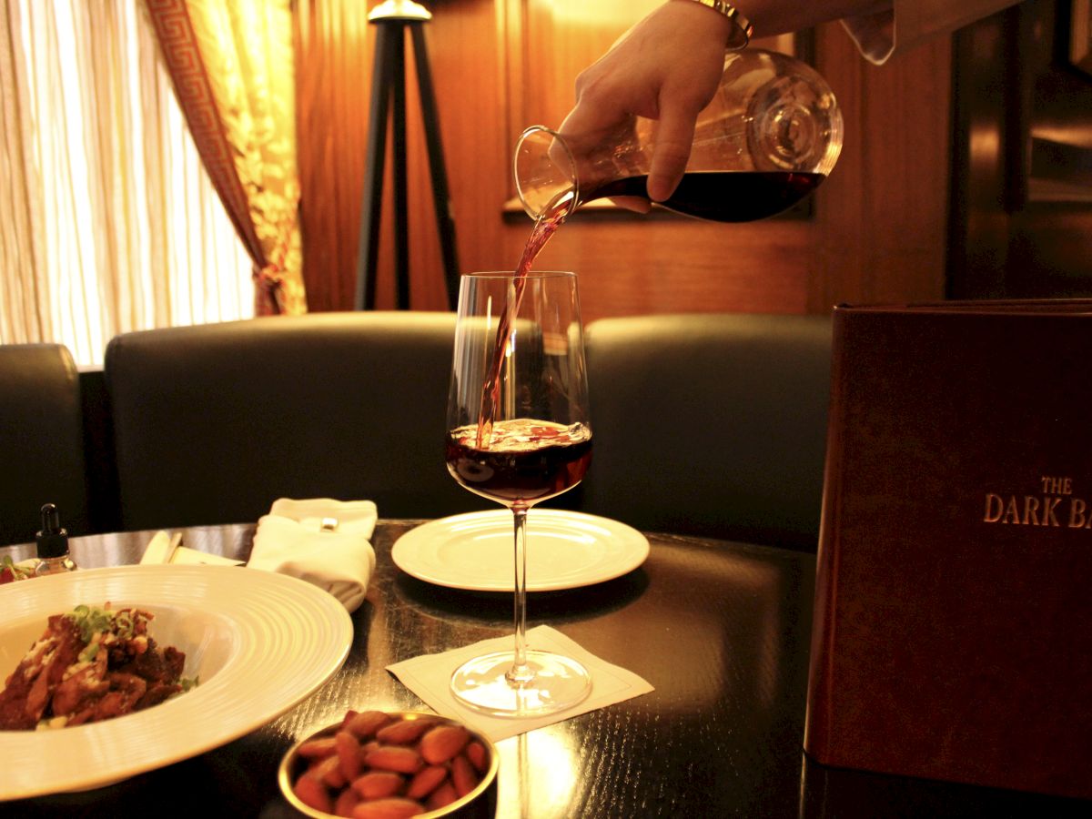 A hand pours red wine into a glass on a table set with dishes, almonds, and a "The Dark Bar" menu in a cozy, dimly lit setting.