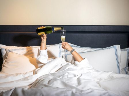 A person in bed is pouring champagne into a glass, surrounded by white bedding and pillows, with only their hands visible.