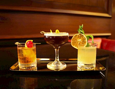 The image shows three different cocktails on a tray: a brown drink with a cherry, a dark drink with a twist, and a light drink with garnishes.