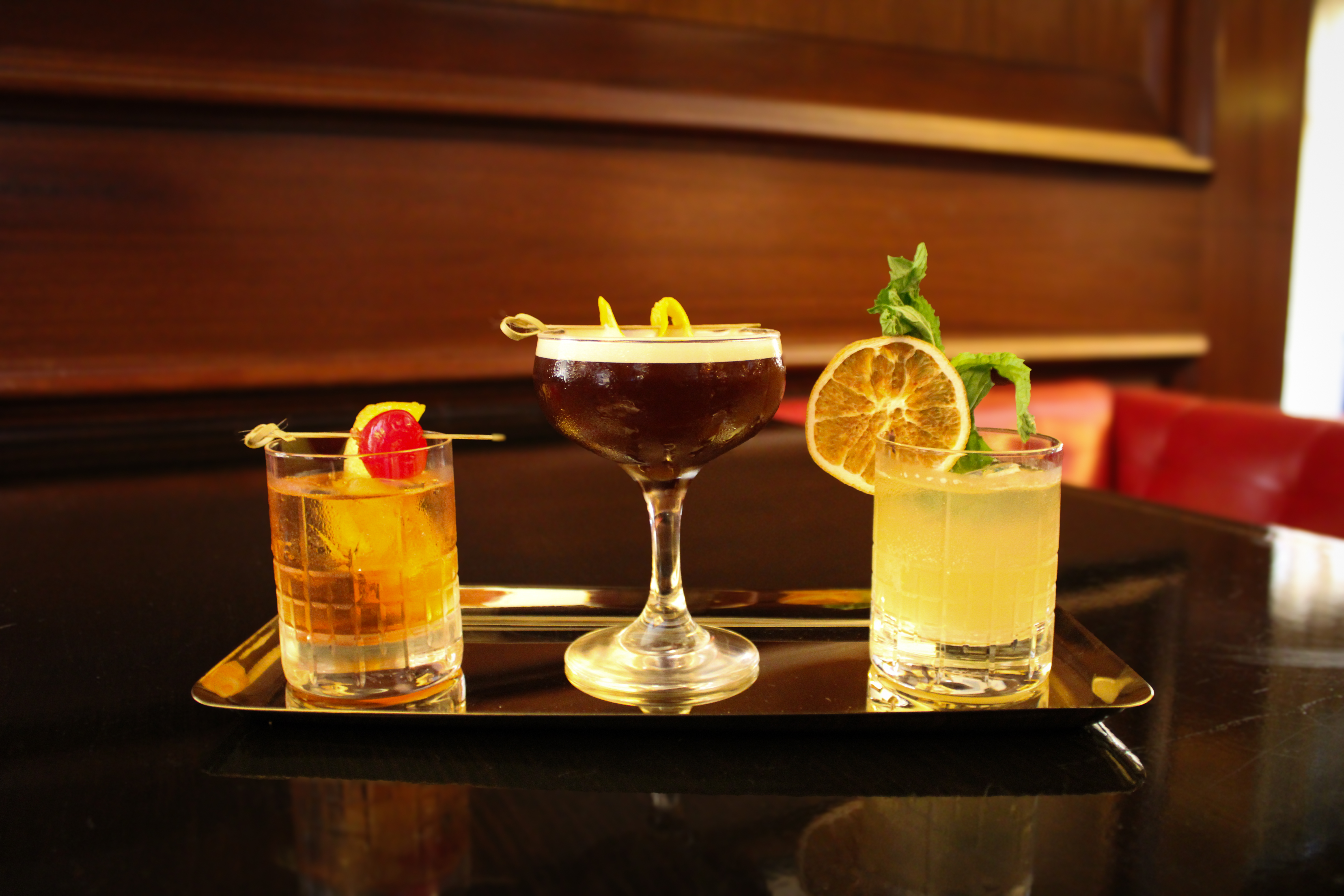 The image shows three different cocktails on a tray: a brown drink with a cherry, a dark drink with a twist, and a light drink with garnishes.