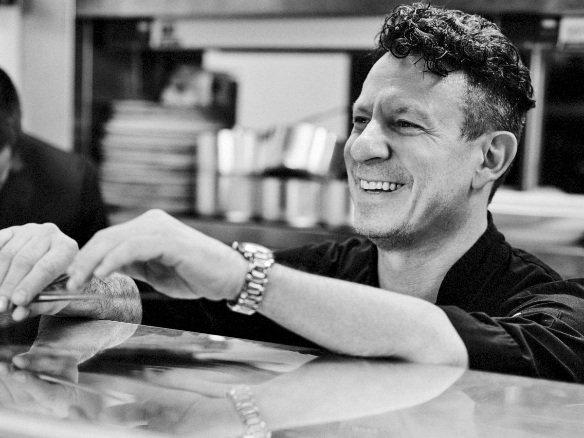 A man smiling, leaning on a counter in a kitchen with blurred shelves and plates in the background, in black and white.