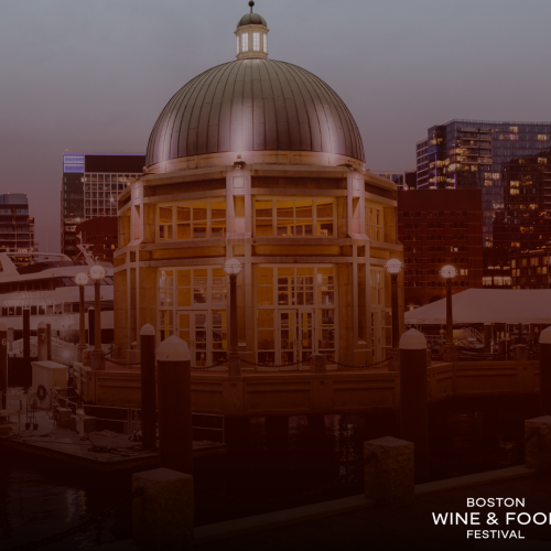 The image shows a waterfront building with a domed roof, illuminated at dusk, with city skyscrapers in the background and text "Boston Wine & Food Festival."