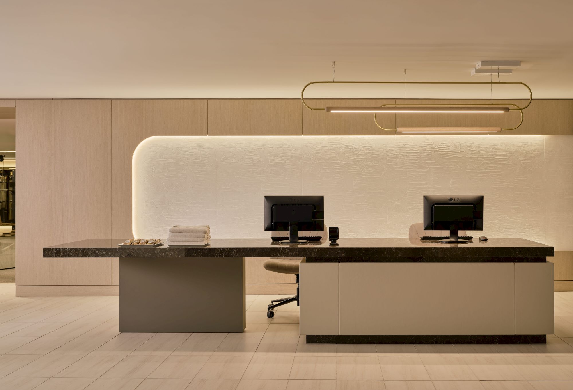 A modern reception desk with two computers, under stylish lighting, set against a minimalist wall design.