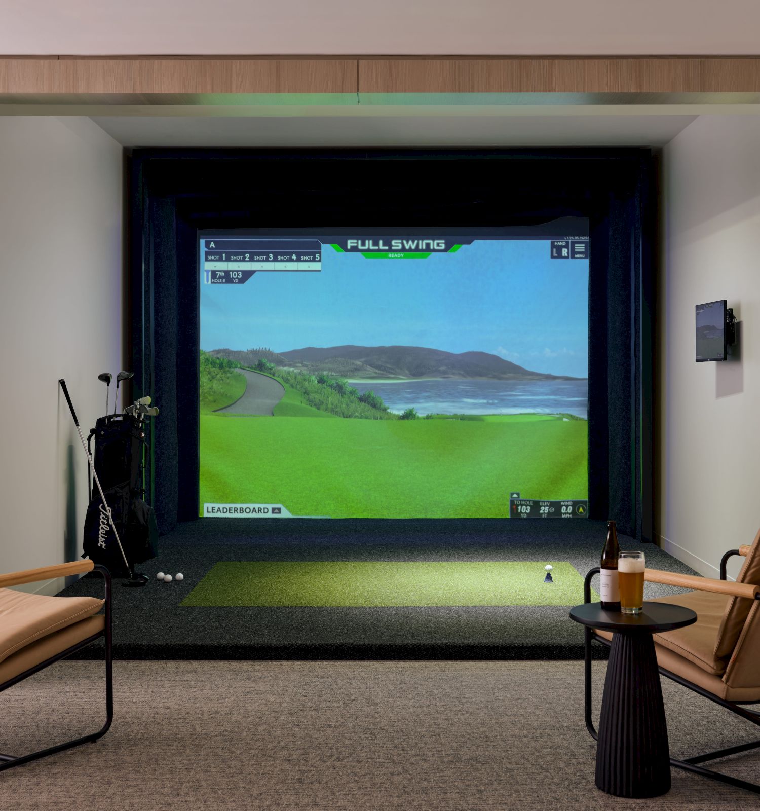 The image shows a golf simulator room with a screen displaying a golf course, two chairs, a side table with a bottle, and golf clubs.