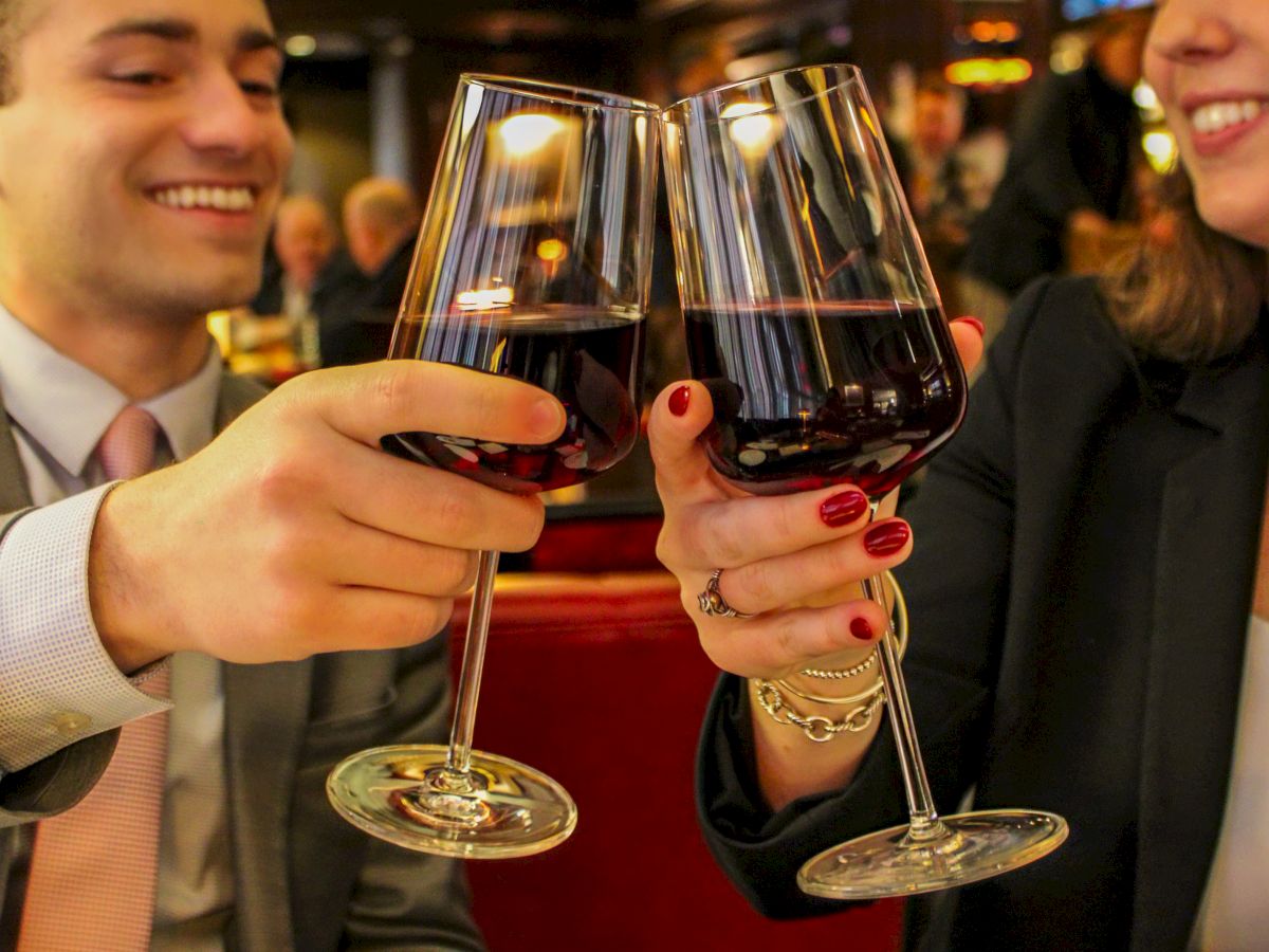 Two people clink glasses of red wine in a celebratory manner at a restaurant, smiling at each other warmly.