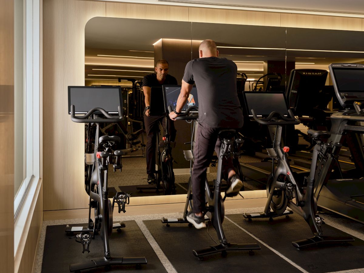 A person on a stationary bike in a gym, reflected in a mirror, with exercise equipment in the background.