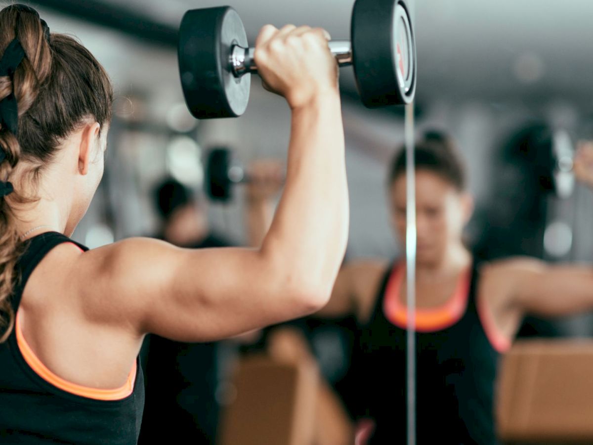 A woman is lifting weights in front of a mirror at the gym, wearing a black tank top with orange accents.