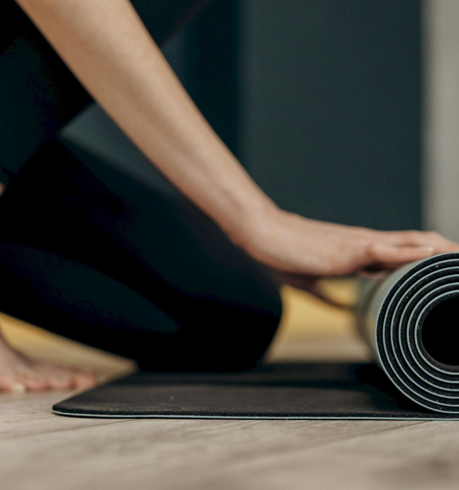 A person is rolling up a black yoga mat on a wooden floor, with their bare feet visible in the background, ending the sentence.