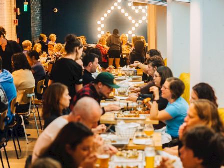 A bustling restaurant scene with people dining, talking, and enjoying their meals at long tables under warm, inviting lighting.