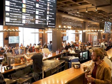 A busy brewery or bar with people socializing, a large menu board displaying drink options, and bartenders serving drinks behind a wooden counter.