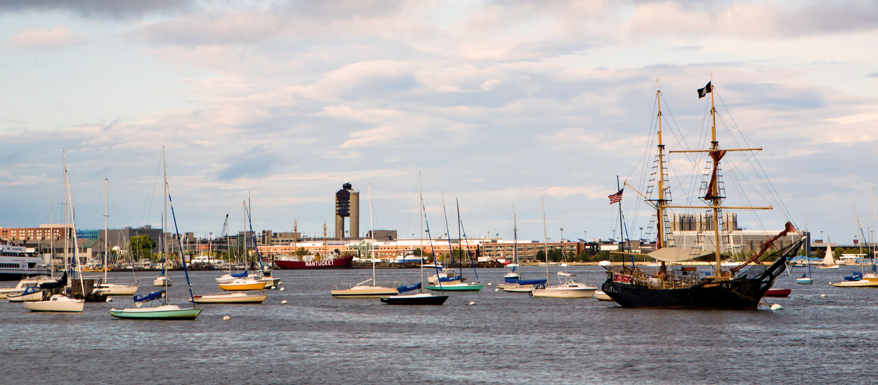 The image shows a harbor with various boats and ships docked, under a sky with some cloud cover.