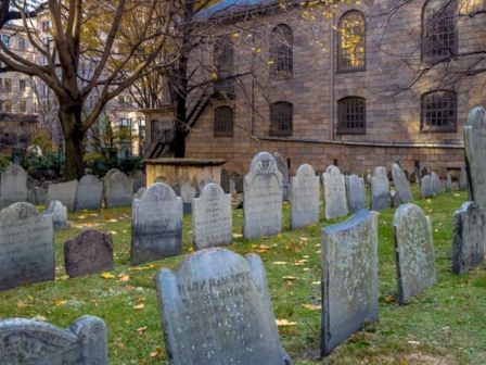 The image shows an old cemetery with numerous tombstones of varying shapes and sizes. A brick building with arched windows is in the background.