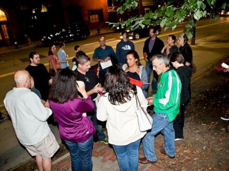 A group of people is gathered outdoors on a sidewalk at night, seemingly engaged in a discussion or activity under a tree.