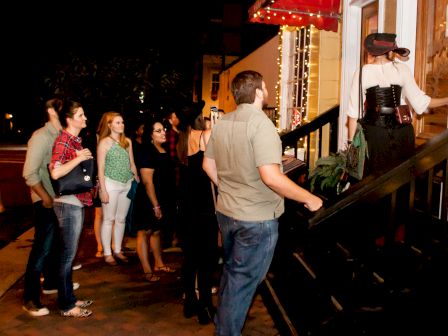 People are standing in line outside of a building, likely waiting to enter, while a person at the front of the line appears to be talking to someone at the door.