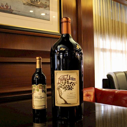 The image shows two wine bottles on a polished wooden surface, one significantly larger than the other, set against a cozy, dimly lit background.