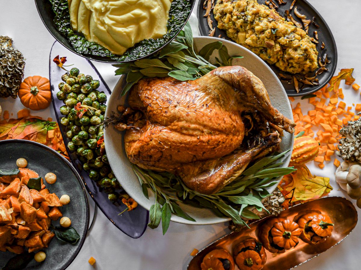 The image shows a Thanksgiving feast with a whole roasted turkey, mashed potatoes, stuffing, sweet potatoes, Brussels sprouts, and decorative pumpkins.