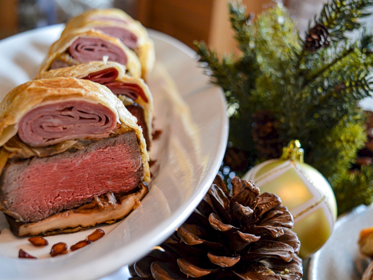 A festive dish featuring slices of Beef Wellington is displayed on a plate, accompanied by decorative pinecones and a Christmas ornament.