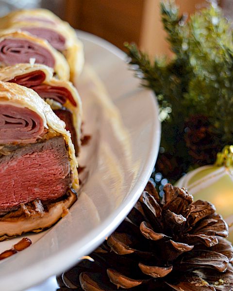 A sliced beef Wellington dish served on a white plate, with holiday decorations including pinecones and ornaments in the background.