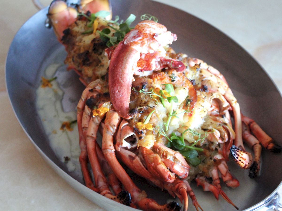 The image shows a cooked lobster served in a metal dish, garnished with greens and melted cheese on top, ready to be eaten.