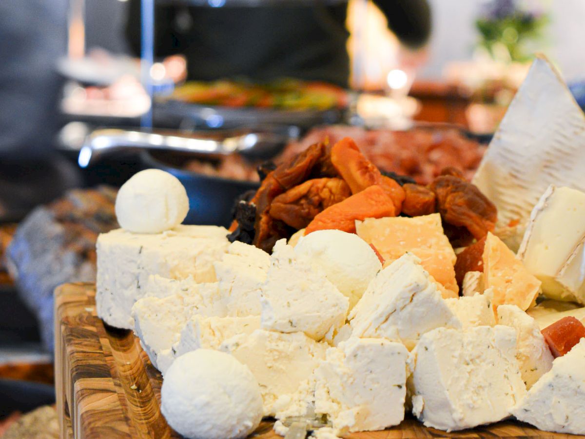 A variety of cheeses and dried fruits are displayed on a wooden board, with more food in the blurred background, creating a gourmet setting.