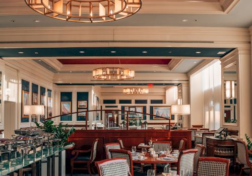 The image shows an elegant restaurant interior with neatly set tables and chairs, large chandeliers, and a warm, inviting ambiance from soft lighting.