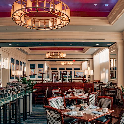 The image shows an elegant restaurant interior with neatly set tables and chairs, large chandeliers, and a warm, inviting ambiance from soft lighting.