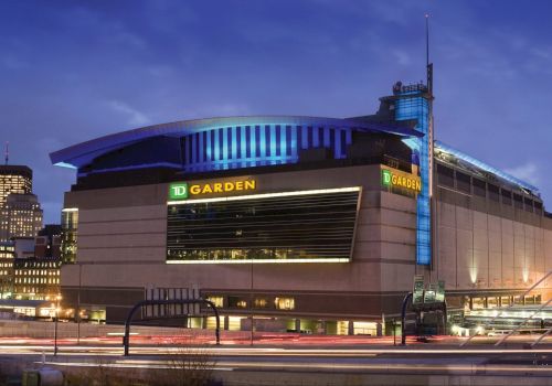 The image shows an exterior view of TD Garden, an arena in a city setting with the skyline illuminated against a twilight sky.